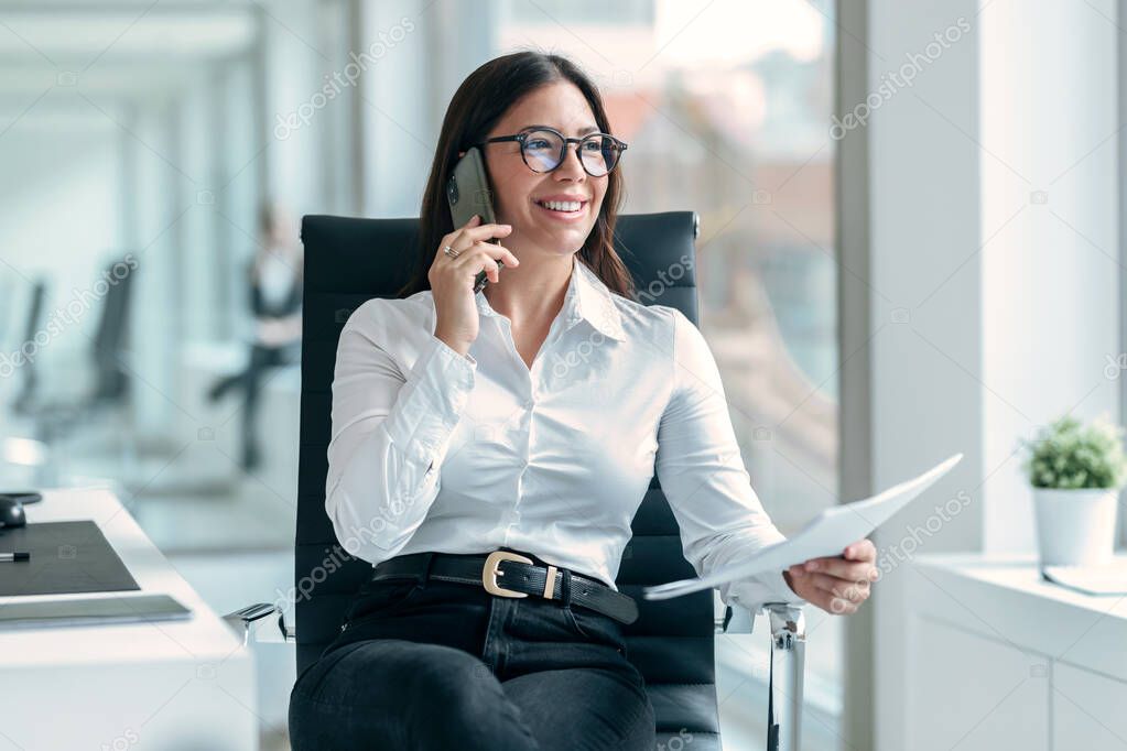 Shot of smiling woman talking with her smartphone while working with computer in the modern startup office.