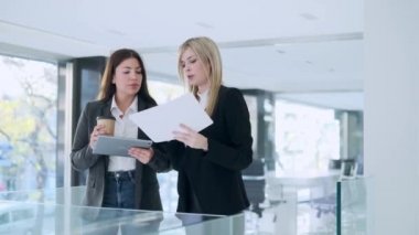 Modern bir girişimde konuşurken ve yürürken dijital tabletle çalışan iki çekici genç iş kadınının videosu. 