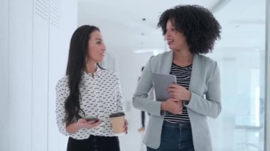 Modern bir girişimde konuşurken ve yürürken dijital tabletle çalışan iki çekici genç iş kadınının videosu. 