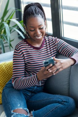 Evdeki kanepede otururken gülümseyen Afrikalı Amerikalı genç bir kadının cep telefonunu kullanması.