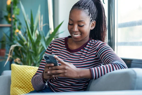 Uśmiechnięta Młoda Afroamerykanka Używająca Telefonu Komórkowego Siedząc Kanapie Domu — Zdjęcie stockowe