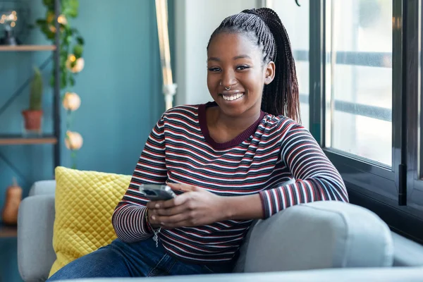 Uśmiechnięta Młoda Afroamerykanka Używająca Telefonu Komórkowego Siedząc Kanapie Domu — Zdjęcie stockowe