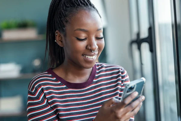 Uśmiechnięta Młoda Afroamerykanka Używająca Telefonu Komórkowego Stojąca Przy Oknie Salonie — Zdjęcie stockowe