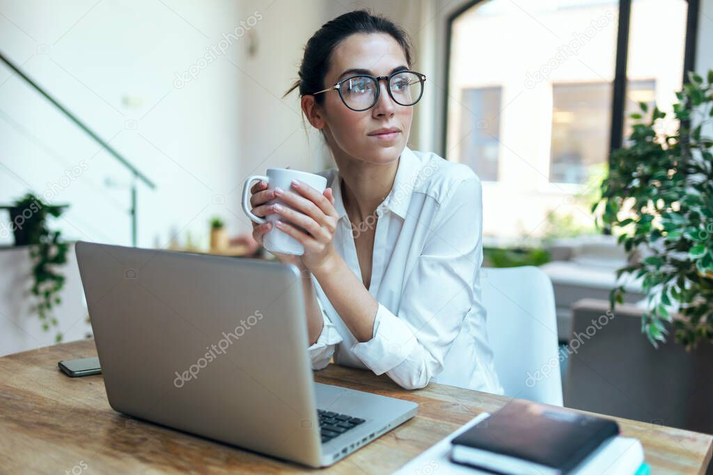 Shot of beautiful woman working with laptop while drinking a cup of coffee looking forward in living room at home.