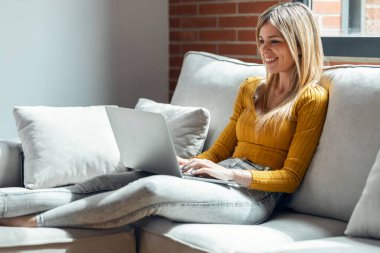 Evdeki kanepede otururken dizüstü bilgisayarıyla çalışan güzel bir genç kadının fotoğrafı..