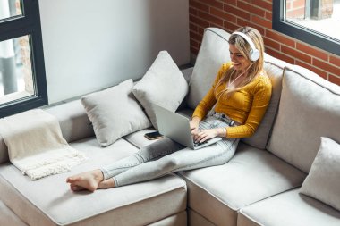 Evdeki bir kanepede oturmuş kulaklıkla müzik dinlerken dizüstü bilgisayarıyla çalışan güzel bir genç kadının fotoğrafı..