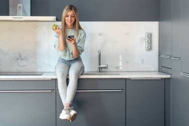 Evdeki mutfakta elma yerken cep telefonunu kullanan güzel bir genç kadının fotoğrafı..