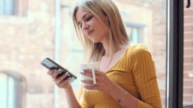 Evdeki oturma odasının penceresinden kahve içerken cep telefonunu kullanan güzel bir kadının videosu..