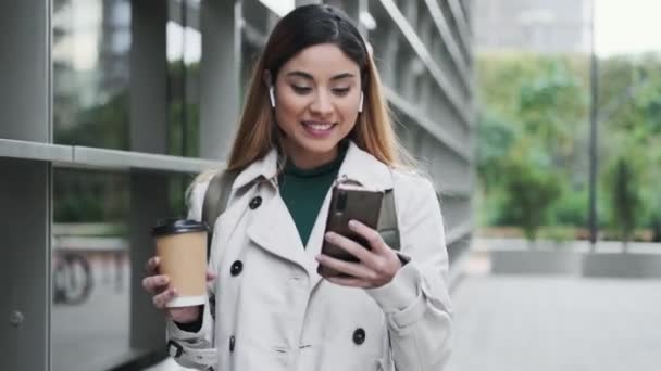 街をカメラを見ながら通りを歩いていると エレガントな女性が音楽に耳を傾け スマートフォンでメッセージを送るビデオ — ストック動画