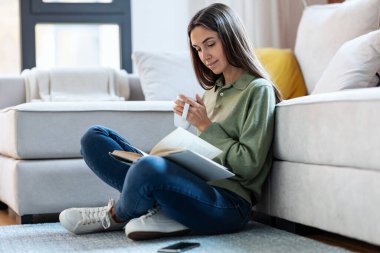 Güzel bir genç kadının kitap okuduğu ve evde yerde otururken bir fincan kahve içtiği bir fotoğraf..