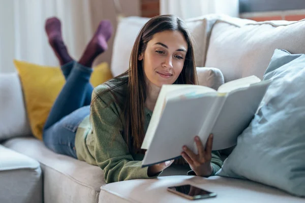 家でソファに横たわっている間に本を読んでいるかなり若い女性のショット — ストック写真