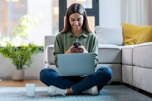 自宅の床に座ってラップトップで作業しながら 彼女の携帯電話を使用してかなり若い女性のショット — ストック写真