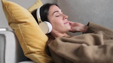 Evdeki kanepede uzanmış akıllı telefon kullanan güzel bir kadının kulaklıkla müzik dinlediği bir video..