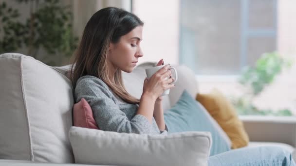 自宅のリビングルームでソファに座っている間にコーヒーを飲みながら美しいです若い女性のビデオ — ストック動画