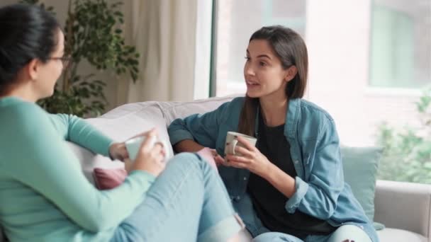二人の笑顔若い女性のビデオを話している間にコーヒーを飲みながらソファに座っている自宅のリビングルーム — ストック動画