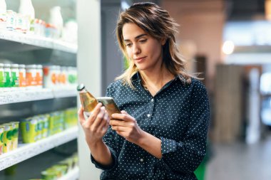 Modern bir kadının markette yürürken ve raftan ürünleri alırken akıllı telefon kullanması.
