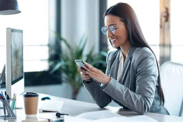 オフィスでコンピュータと仕事をしながら携帯電話を使って笑顔のビジネス女性のショット — ストック写真