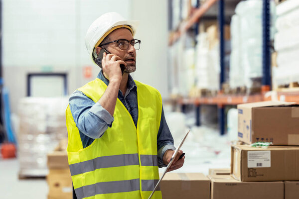 Shot of mature worker talking with smartphone while passing list of material that has arrived at the store warehouse.