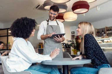 Shot of handsome smiling waitress using application on digital tablet computer to order what the two girls want for breakfast in a pastry shop.