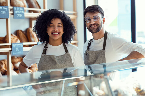 Portrait of two younger owners selling fresh pastry and loaves un bread section and smiling at pastry shop.