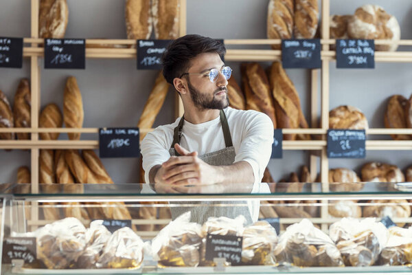 Portrait of handsome younger owner selling fresh pastry and loaves un bread section and smiling at pastry shop.