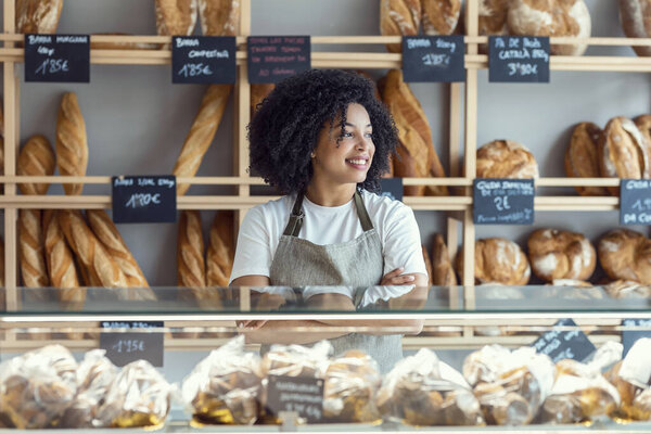 Portrait of beautiful afro younger owner woman selling fresh pastry and loaves un bread section and smiling at pastry shop.