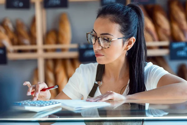 Shot of beautiful woman seller checking stock of pastry while counting the benefits of the day with the calculator in the pastry shop.