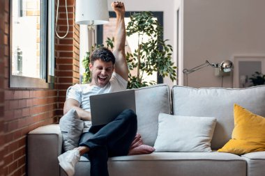 Shot of successful young man using her laptop and celebrating something while sitting on sofa at home.