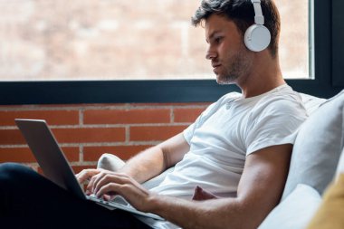 Shot of smart man working with her laptop while listening music with headphones sitting on a couch at home