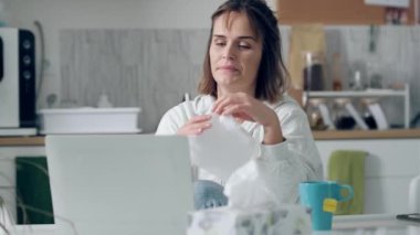 Video of llness young woman sneezing in a tissue while working with laptop sitting in the kitchen at home.