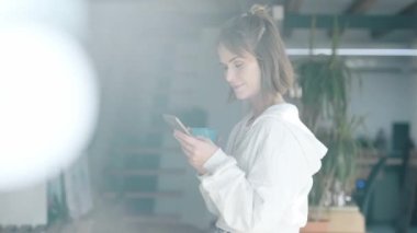 Neşeli bir kadının kahve içerken evinin penceresinin yanında cep telefonu kullanmasının videosu..