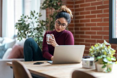 Evdeki oturma odasında kahve içerken bilgisayarla çalışan güzel bir iş kadınının fotoğrafı..