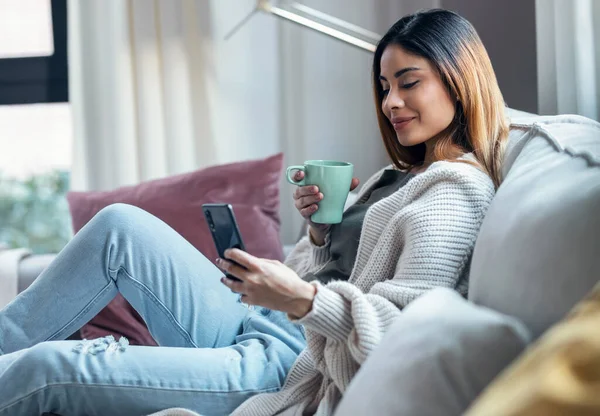 Shot of confident woman using her mobile phone while drinking coffee sitting on sofa at home.