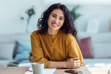Evde oturma odasında kameraya bakarken kendine güvenen bir kadının laptopuyla görüntüsü..