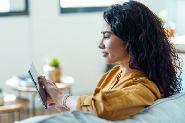 Evdeki kanepede otururken dijital tabletini okumak için kullanan güzel bir genç kadının fotoğrafı..