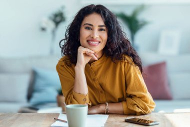 Evde oturma odasında kameraya bakarken kendine güvenen bir kadının laptopuyla görüntüsü..