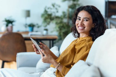 Evdeki kanepede otururken dijital tabletini okumak için kullanan güzel bir genç kadının fotoğrafı..