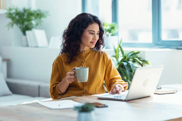 自宅のリビングルームでコーヒーを飲みながらノートパソコンで働く集中美しいビジネス女性のショット — ストック写真