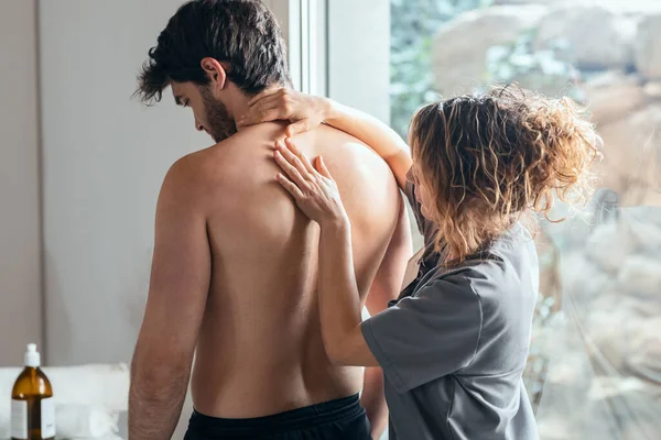 診療所でリハビリテーション療法の痛みをしながら 男性患者の背中を負傷した治療を検討する女性理学療法士の仕事のショット — ストック写真