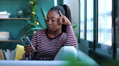 Evdeki kanepede otururken cep telefonunu kullanan neşeli Afro-Amerikan kadının videosu..