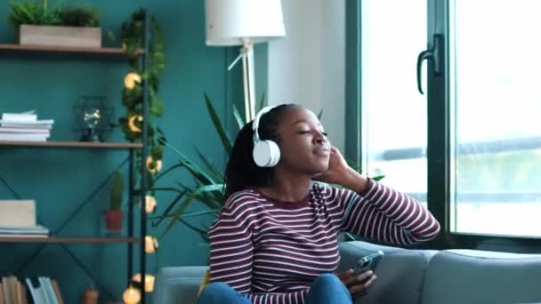 自宅でソファに座っているスマートフォンを使用している間に音楽を聞いて美しい女性のビデオ — ストック動画