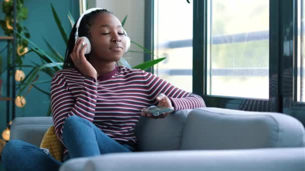 自宅でソファに座っているスマートフォンを使用している間に音楽を聞いて美しい女性のビデオ — ストック動画
