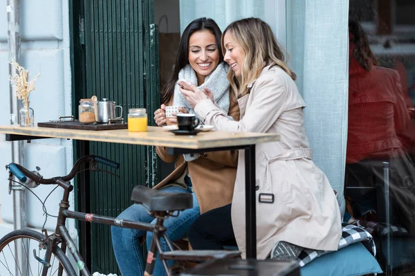 Shot of lovely girls couple sharing a brunch together while talking and looking the smartphone on the healthy coffee shop terrace.
