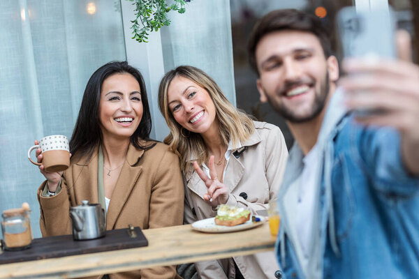 Shot of group of cool friends sharing a brunch together while taking a selfie with smartphone on the healthy coffee shop terrace.