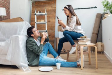 Yeni evlerindeki karton kutularda akıllı telefonlu komik fotoğraflar çekerken sevimli bir çiftin sandviç yemesi.