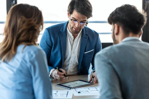 Shot of handsome real-estate agent showing house plans on electronic tablet while talking to the couple about buying the house in the office.