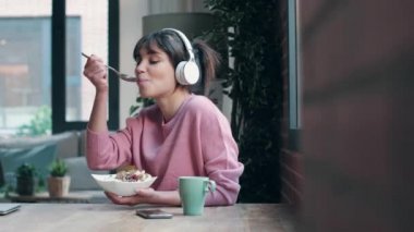 Video of beautiful young woman listening music with headphones while having healthy breakfast in the living room at home.