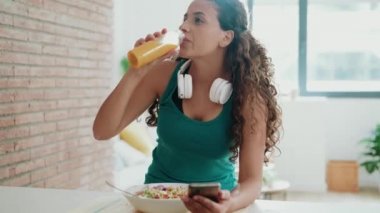 Video of beautiful sporty woman eating a bowl of salad while messaging with her mobile phone in the kitchen at home.