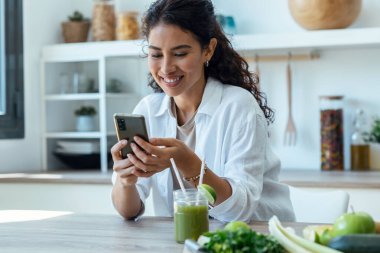 Shot of pretty woman using her mobile phone while drinking fruit detox juice in the kitchen at home.