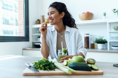 Shot of pretty woman eating a piece of cucumber while drinking fruit detox juice in the kitchen at home.
